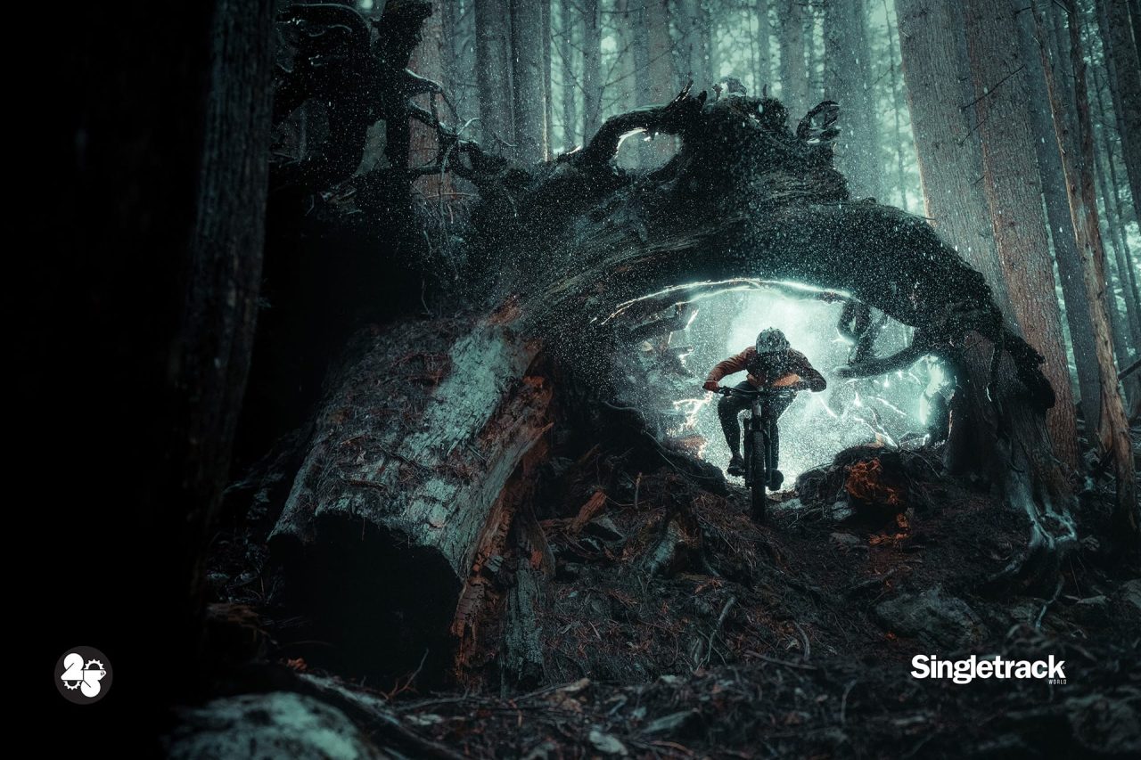 A mountain biker rides through a large fallen tree in a dark, misty forest, creating a dramatic scene with splashes of water and debris.