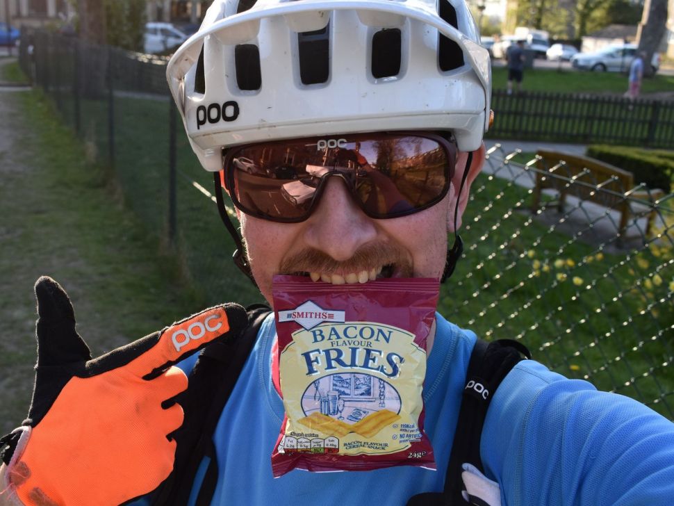 A cyclist wearing a helmet and sunglasses is smiling and holding a bag of bacon-flavored fries while posing for the camera.