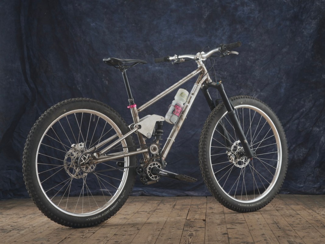 A custom-built full suspension mountain bike featuring adjustable geometry, a pinion gearbox, and mixed wheel sizes (27.5 inch rear and 29 inch front). The bike is shown against a dark textured background.