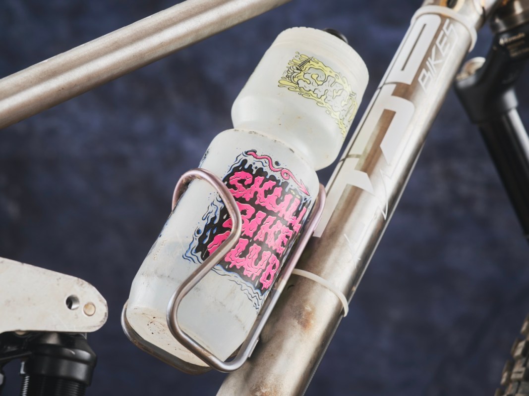A close-up of a bicycle frame featuring a water bottle holder with a 'Skull Bike Club' sticker on the bottle, showcasing a metallic frame structure.