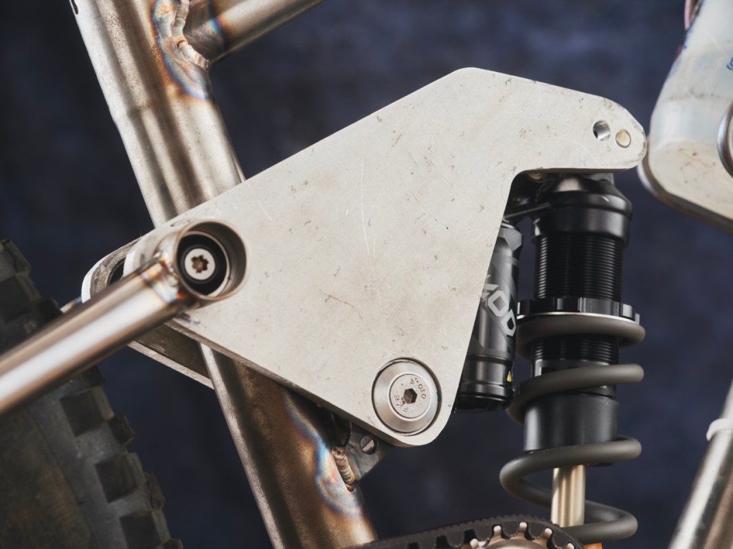 Close-up of a custom mountain bike suspension linkage system, featuring a metallic linkage plate and a shock absorber, showcasing precise welding and design elements.