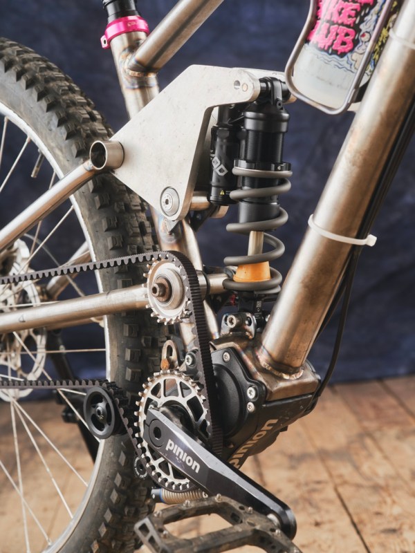 Close-up of a custom full-suspension mountain bike showing the pinion gearbox, shock absorber, and belt drive system, with a focus on the adjustable linkage for suspension tuning.