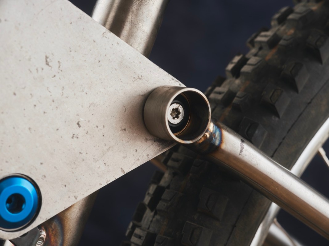 Close-up view of a bicycle frame component, featuring a metallic linkage with a circular socket and blue detailing, positioned near a textured tire.
