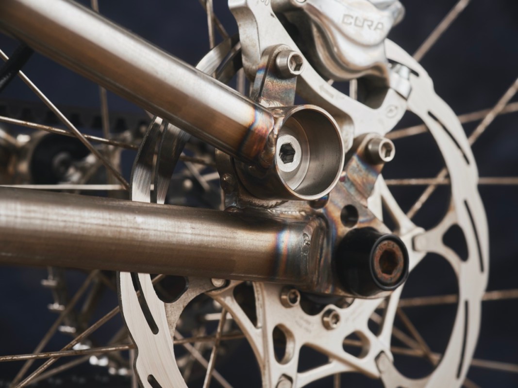 Close-up of a bicycle's rear suspension linkage and brake disc, showcasing the intricate welding details and metallic finish.