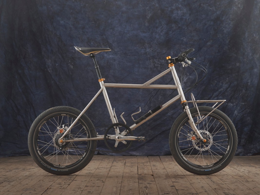 A custom-built, adjustable mountain bike featuring a stainless steel frame, Pinion gearbox, and mixed wheel sizes. The bike is shown on a wooden floor with a dark blue backdrop.