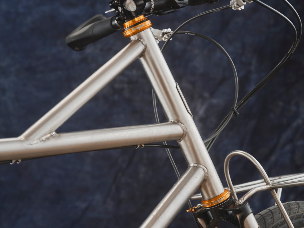 Close-up of a bicycle frame showcasing silver metal tubing and an orange headset, with visible welding details against a dark background.