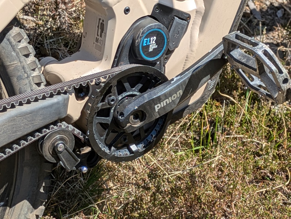 Close-up of the Pinion gearbox system and crankset on a Moustache e-bike, showcasing the carbon belt drive and gears against a natural, grassy background.