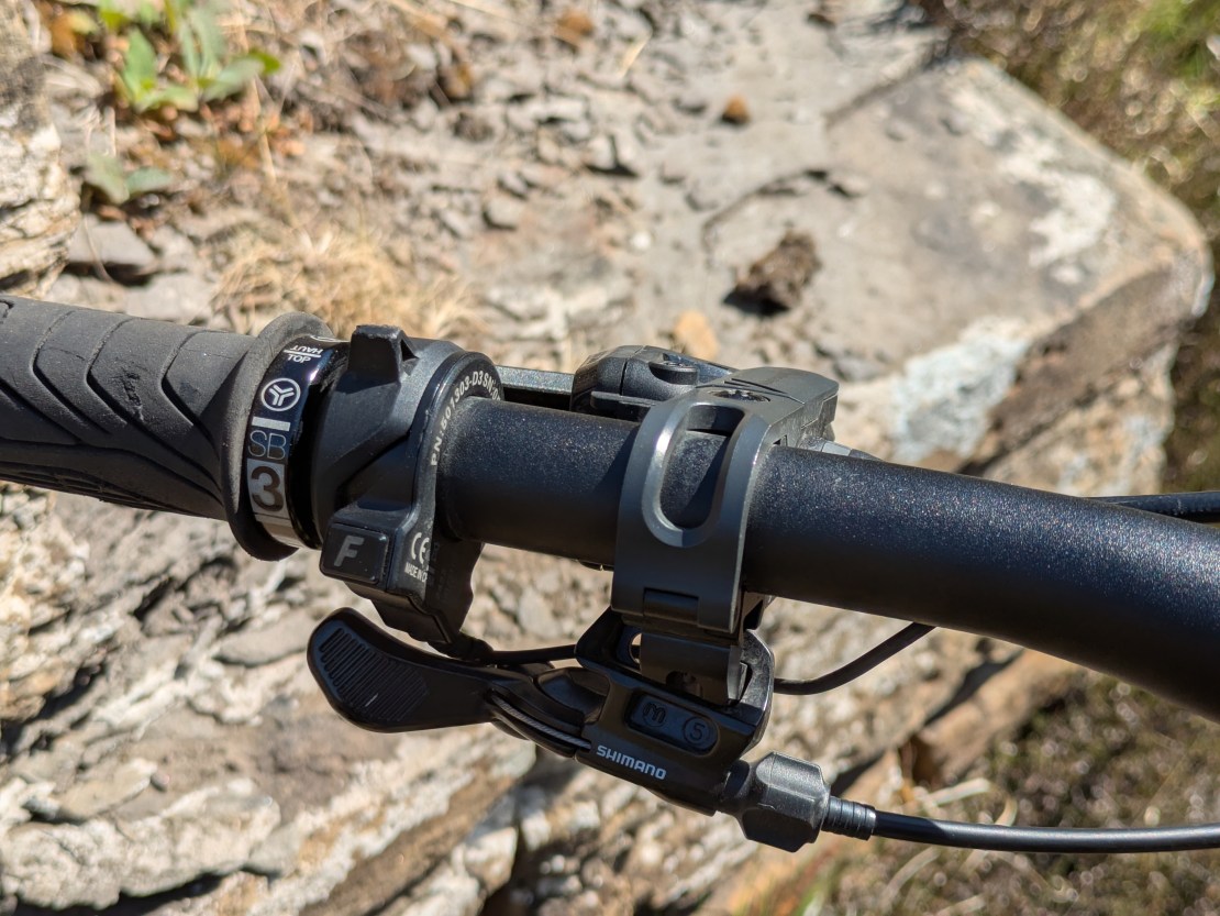 Close-up view of bike handlebar controls, featuring a gear shift lever and remote display mount against a rocky surface.