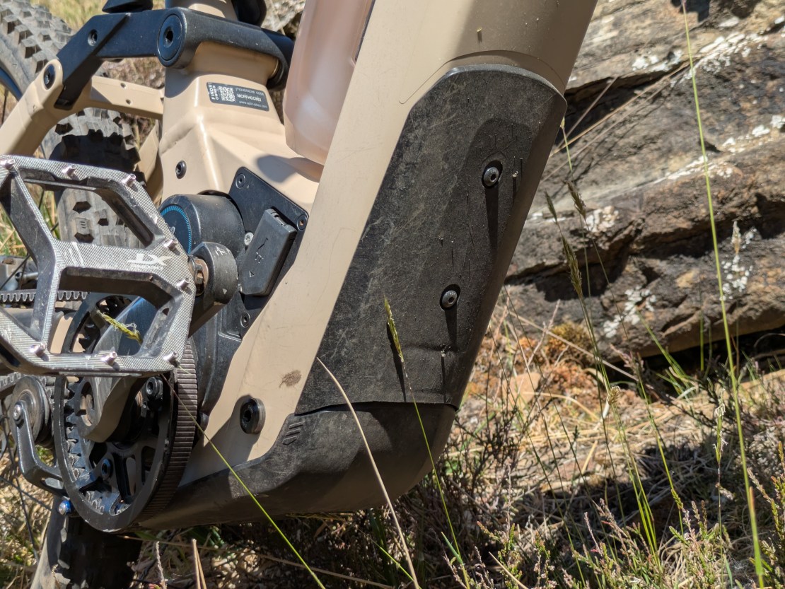 Close-up view of the drivetrain and gearbox of the Moustache Clutch electric mountain bike, showcasing the Pinion MGU motor, chainring, and protective cover against a rocky background.