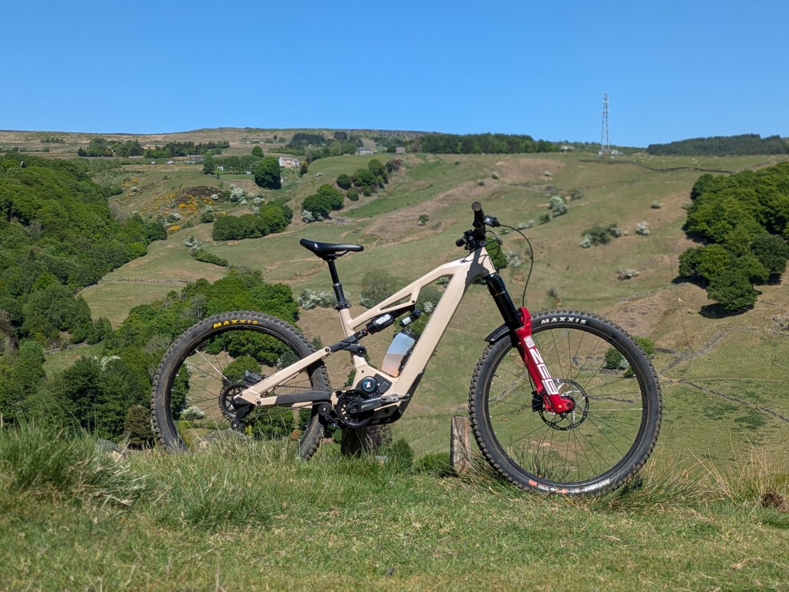A Moustache electric mountain bike with a Pinion MGU gearbox motor, positioned on a grassy hillside with verdant greenery and clear blue skies in the background.