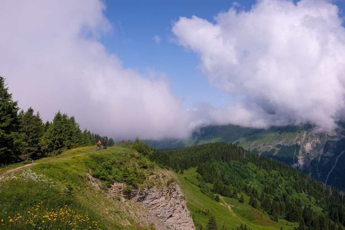 A mountain biker riding along a scenic trail with lush greenery and clouds in the background, near Morzine, France.