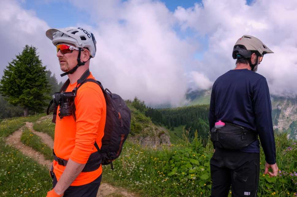 Two mountain bikers wearing helmets and bright clothing stand on a trail overlooking a scenic landscape with trees and clouds in the background.