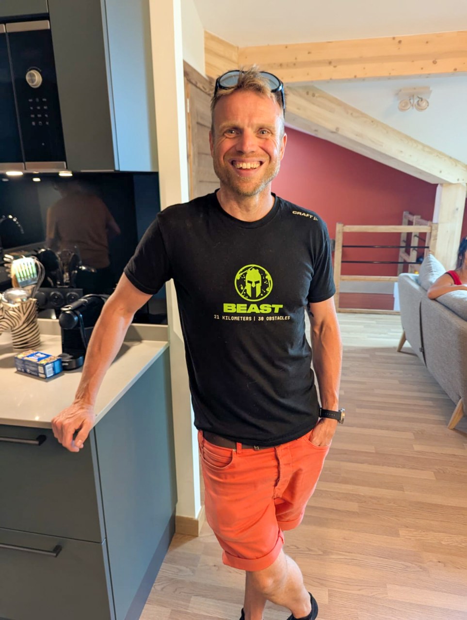 A smiling man standing in a modern kitchen, wearing a black t-shirt with a green logo and orange shorts.