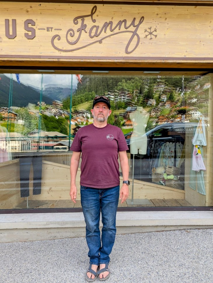 A man standing outside a shop with a wooden facade and a sign that reads 'BUS ET Fanny'. The background shows a view of green hills and buildings.