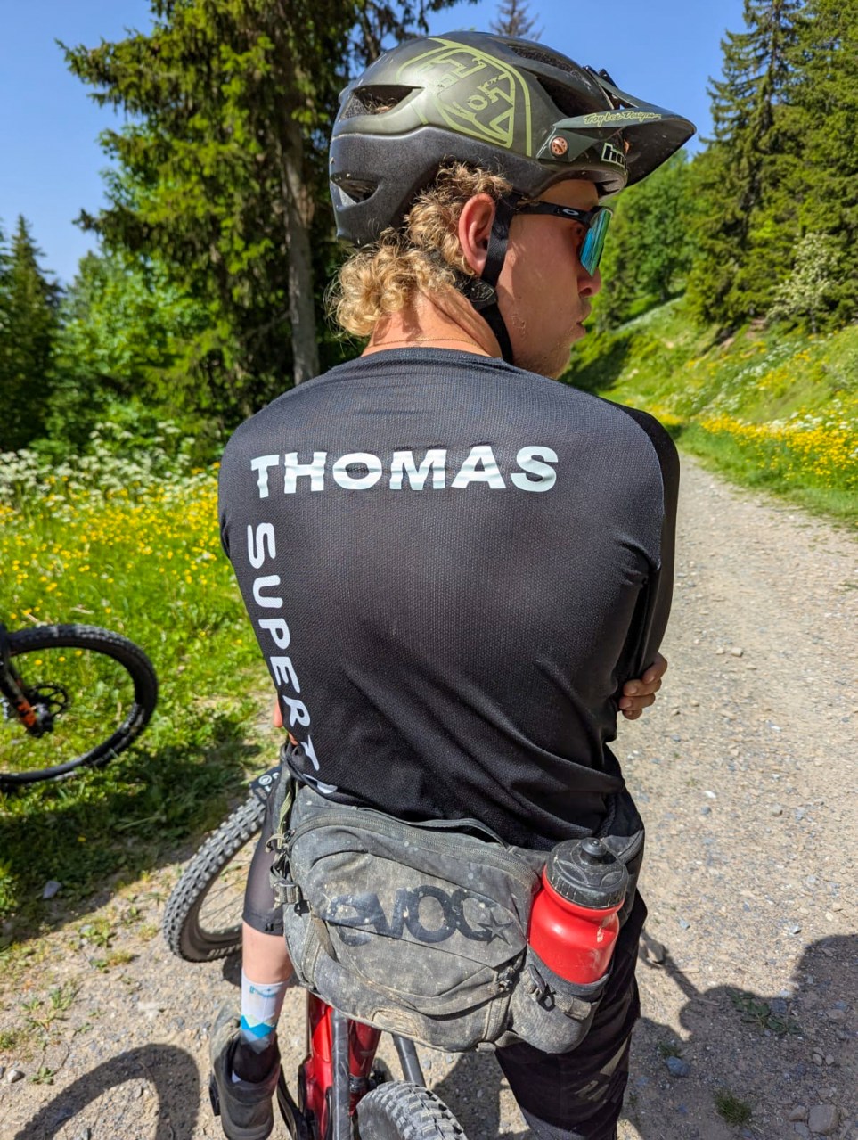 A mountain biker wearing sunglasses and a helmet with the name 'THOMAS' on their back, resting on a trail surrounded by trees and wildflowers.