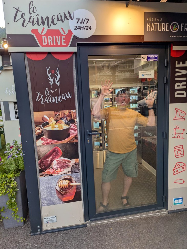 A person standing inside a glass-fronted kiosk named 'le traîneau DRIVE', with various food items displayed on the signage, in a scenic outdoor location.