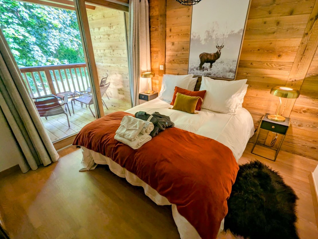 Cozy bedroom with a large bed, decorative pillows, and a warm blanket, featuring a wooden wall and balcony access overlooking greenery. Bike Morzine