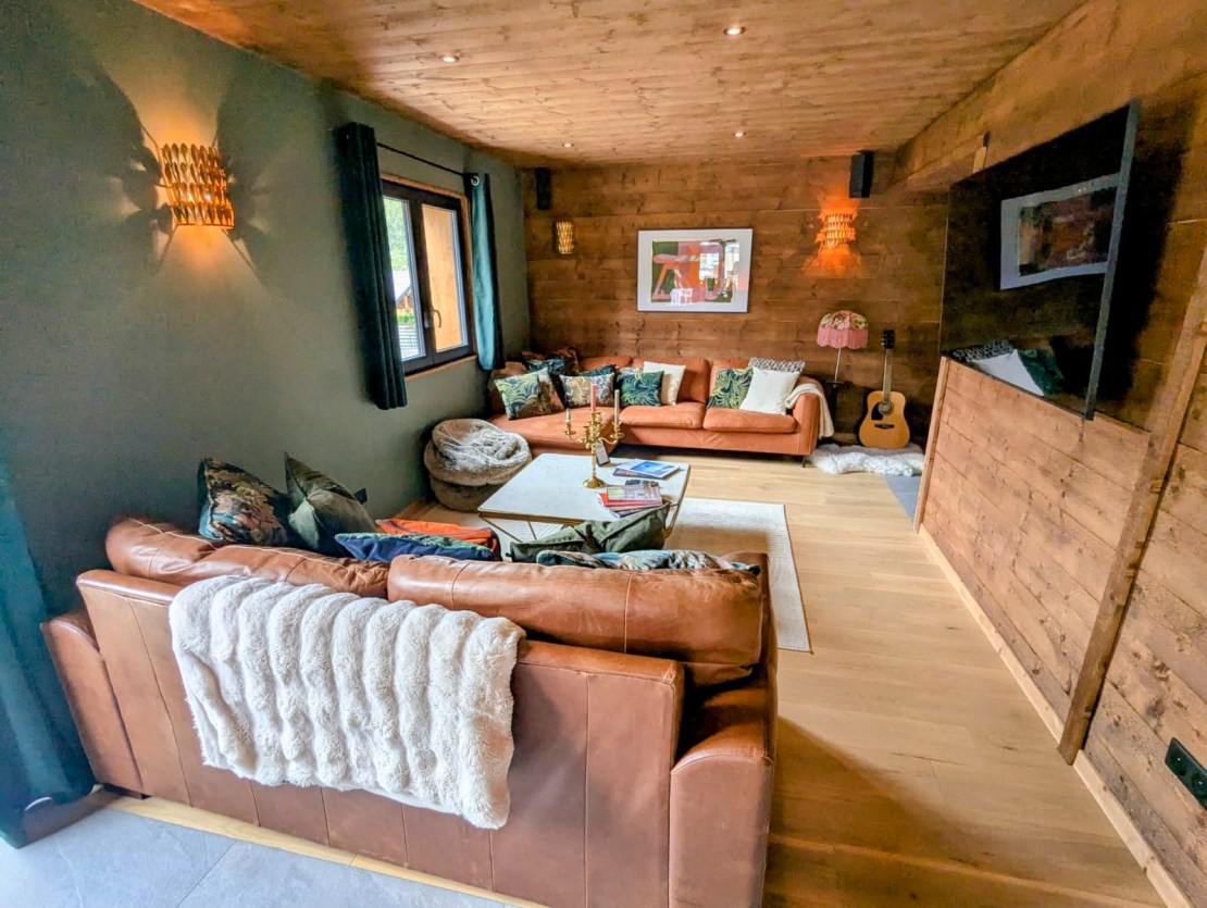 Cozy living room with brown leather sofas, wooden walls, and decorative pillows, featuring a coffee table and a guitar in the corner. Bike Morzine