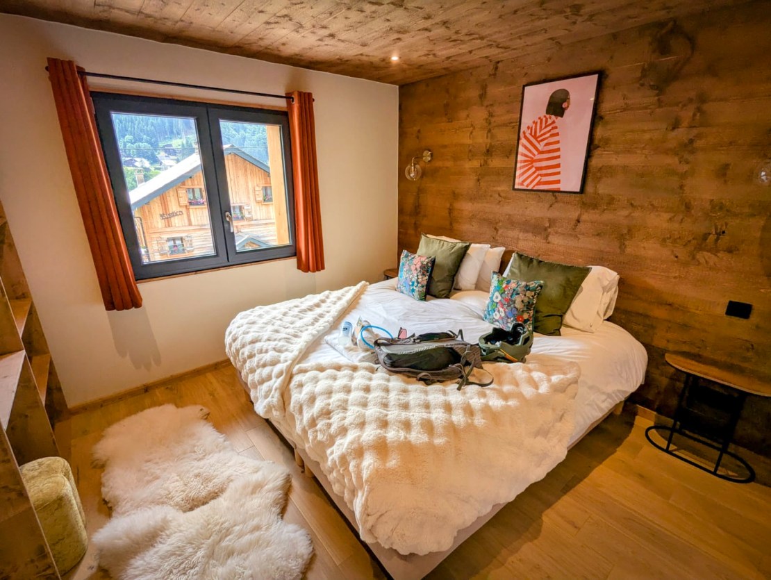 Cozy bedroom with a large bed, decorative pillows, and a warm color scheme, featuring wooden walls and a window overlooking a mountain village. Bike Morzine