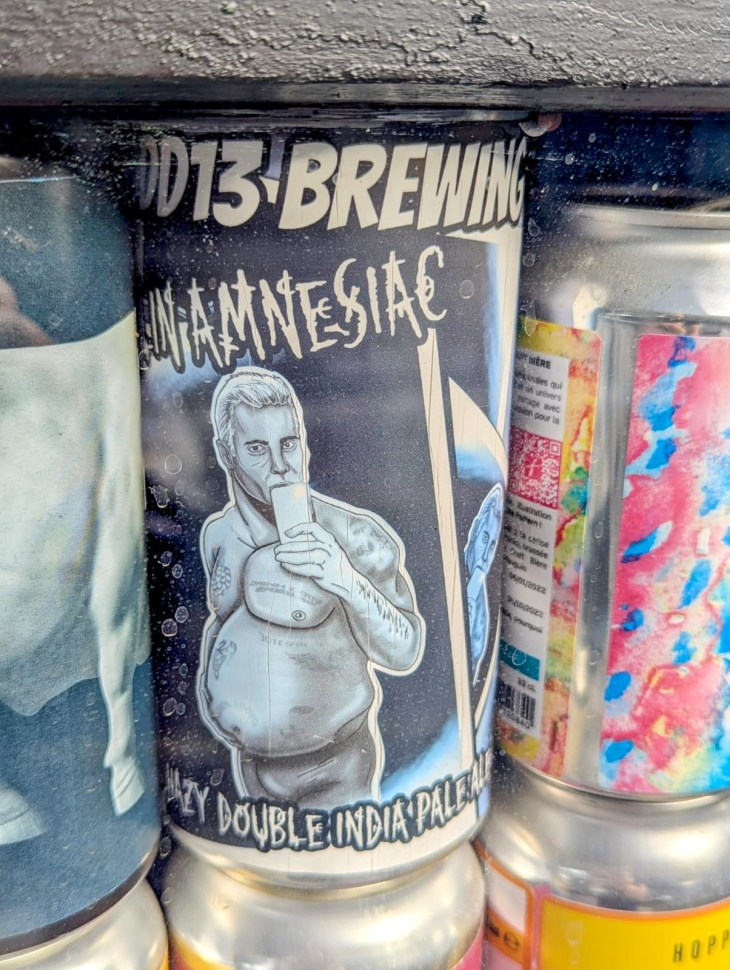 A close-up of a beer can from D13 Brewing featuring a cartoonish depiction of a man holding a phone in front of his chest. The can is labeled 'Anamnesiac' and indicates it is a hazy double India pale ale.