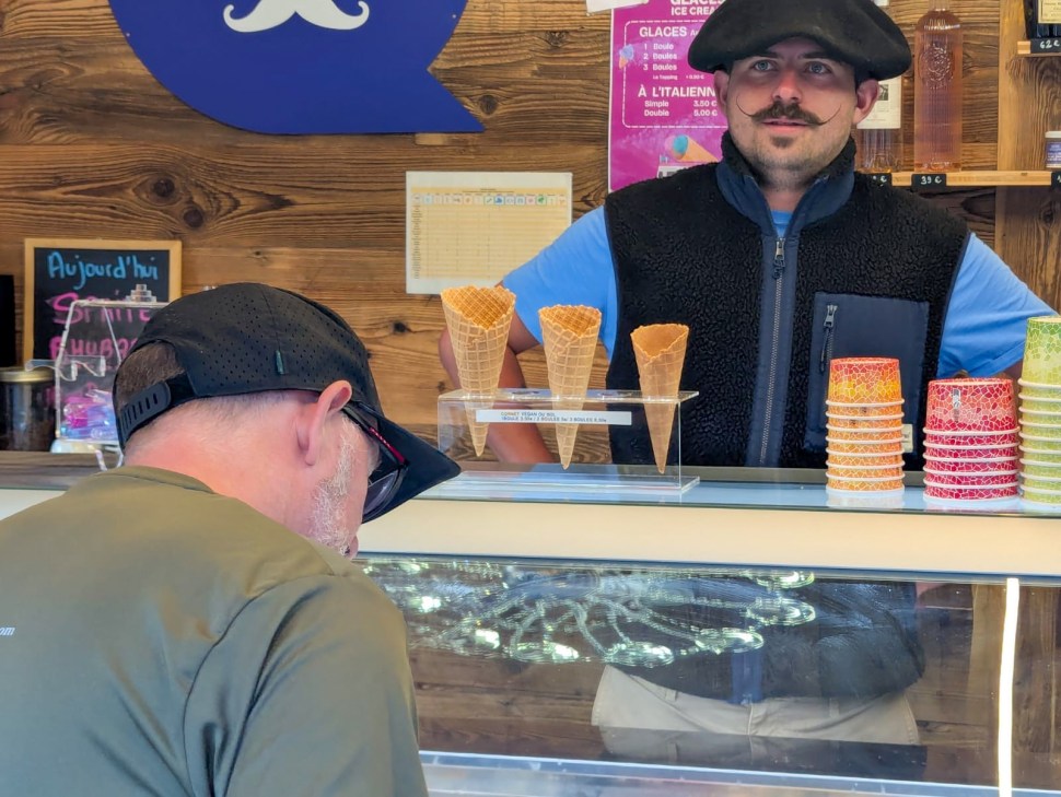 A man with a mustache and beret stands behind an ice cream counter, serving a customer who is turned away, focused on the selection of ice cream cones.