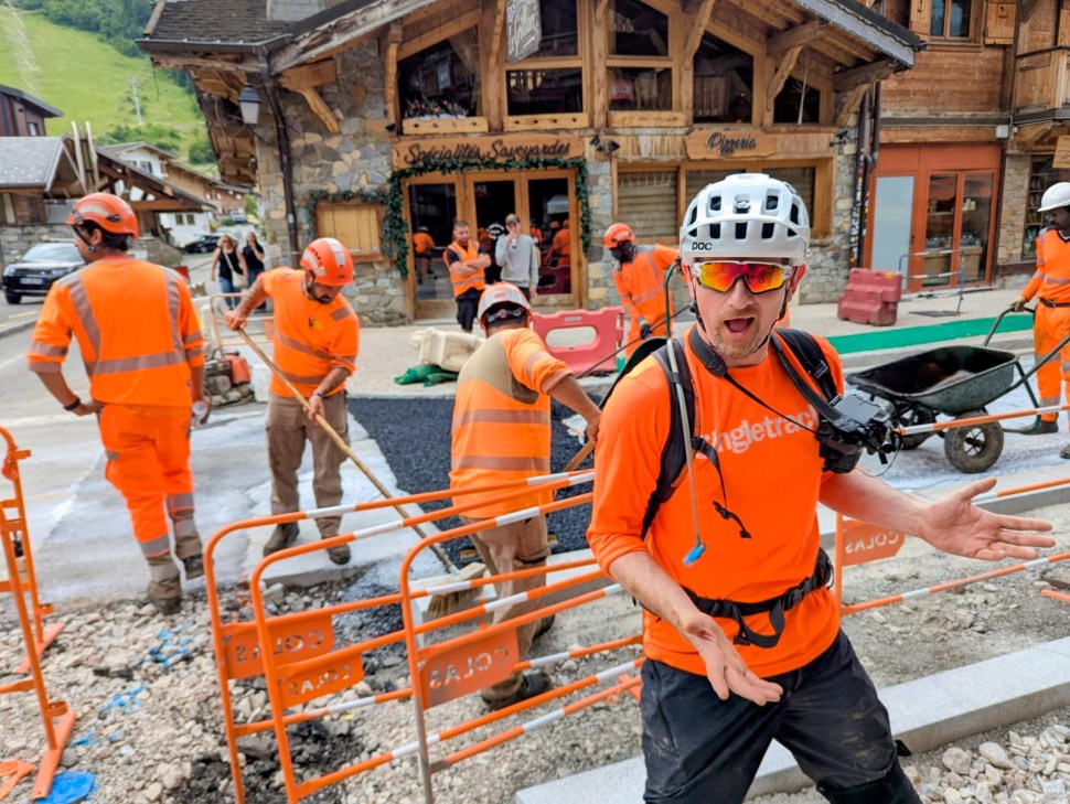 Construction workers in orange uniforms work on a street, with a group of onlookers in the background.