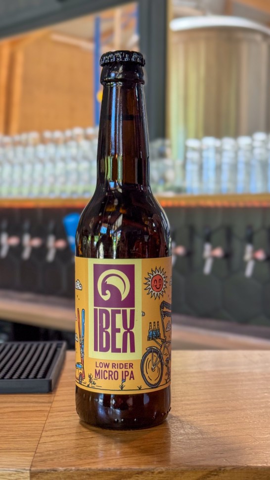 Bottle of Ibex Low Rider Micro IPA beer on a wooden bar top with blurred background.
