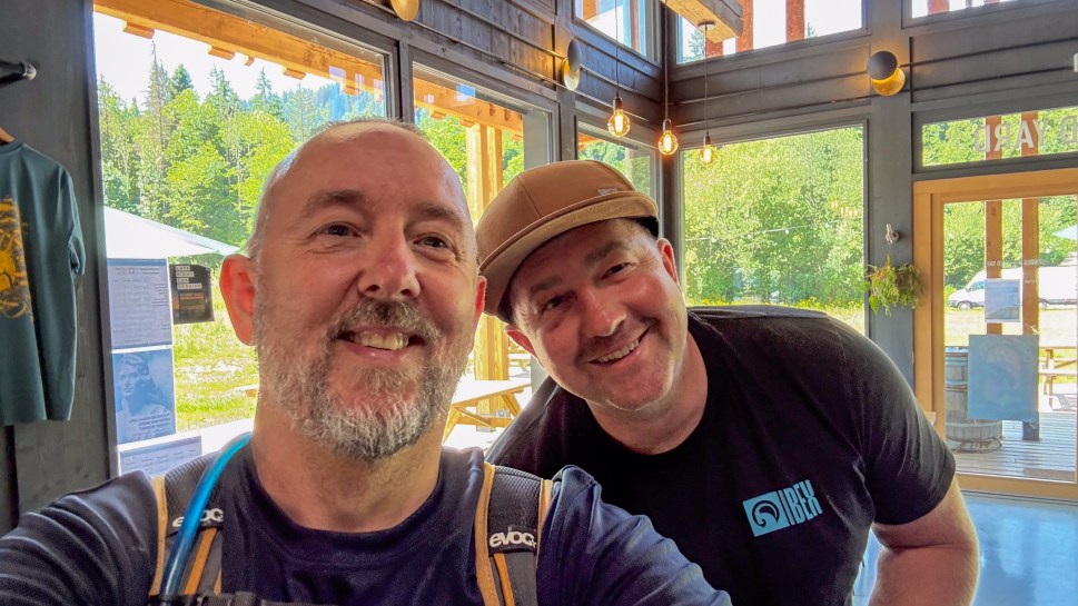 Two men smiling for a selfie inside a modern building with large windows and natural lighting.