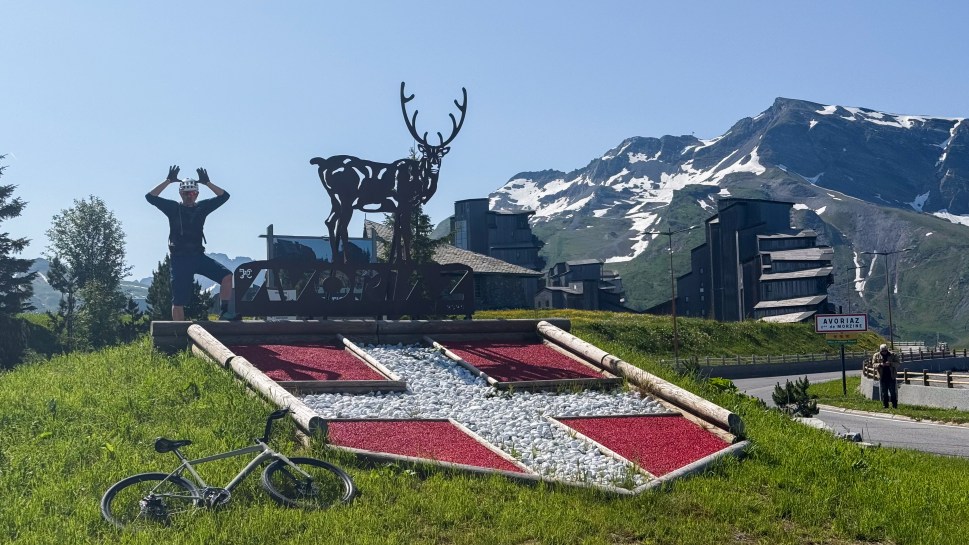 A cyclist posing near a sign for Avoriaz, surrounded by green grass and mountains in the background. A bike is resting on the ground nearby.