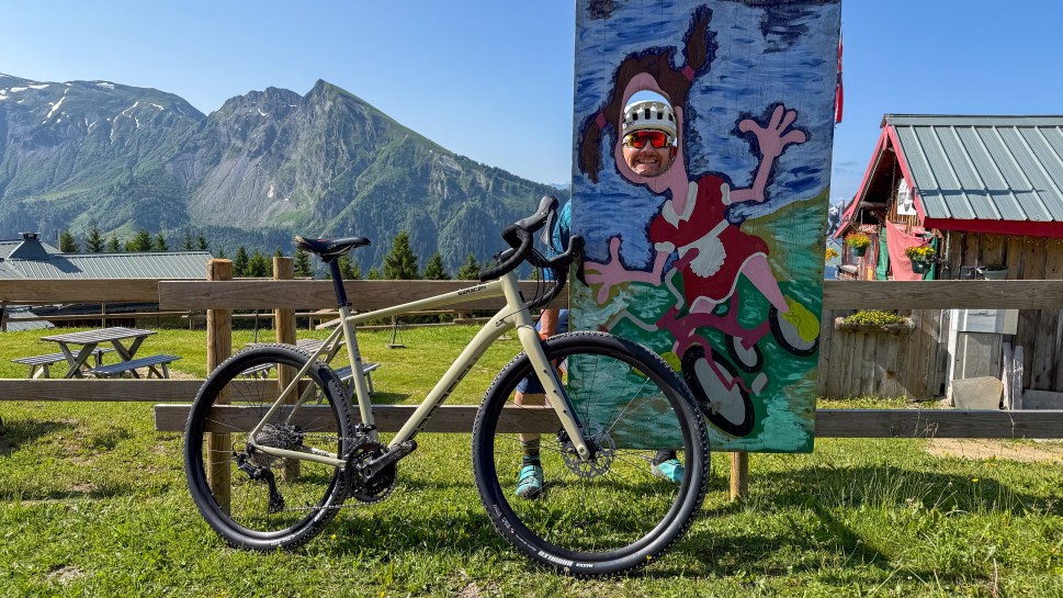 A mountain bike propped against a wooden fence, with a painted cutout of a cyclist behind. Scenic mountains and a clear blue sky are in the background.
