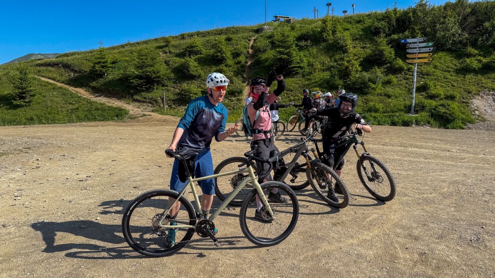 A group of mountain bikers posing and celebrating at a trailhead in Morzine, France, with green hills and a signpost in the background.