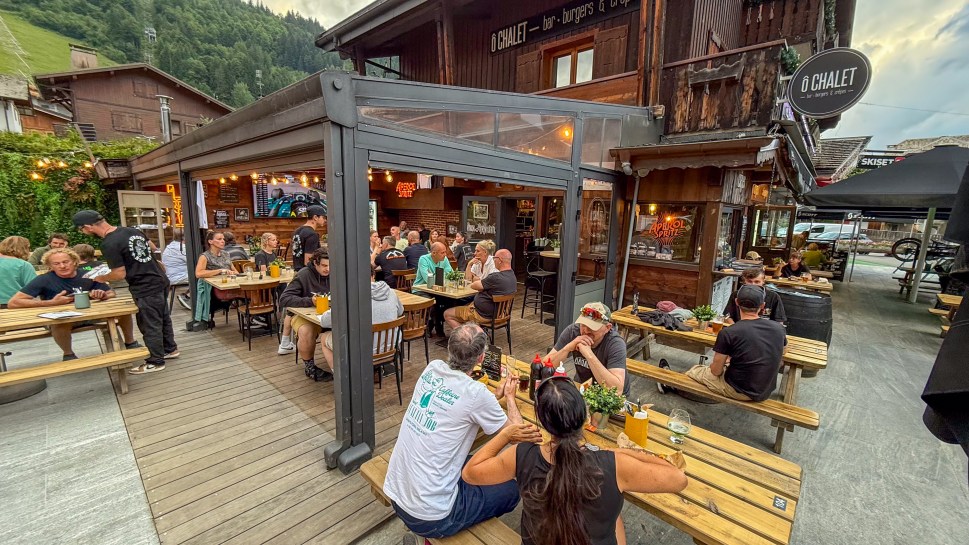 Outdoor dining area of O'Chalet restaurant in Morzine, France, filled with guests enjoying their meals and drinks surrounded by wooden structures and greenery.
