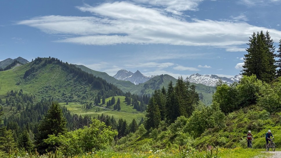 Two mountain bikers riding on a trail surrounded by lush green hills and pine trees, with distant snow-capped peaks under a partly cloudy sky.