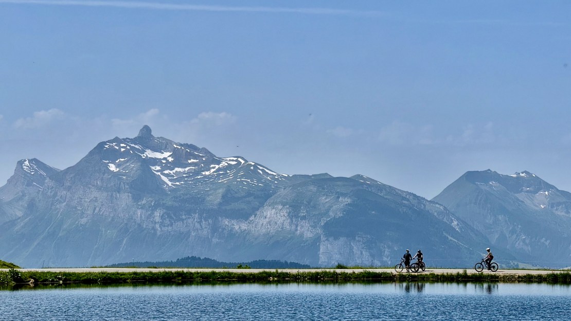 Bicyclists riding along a path by a lake with mountains in the background in Les Gets, France.