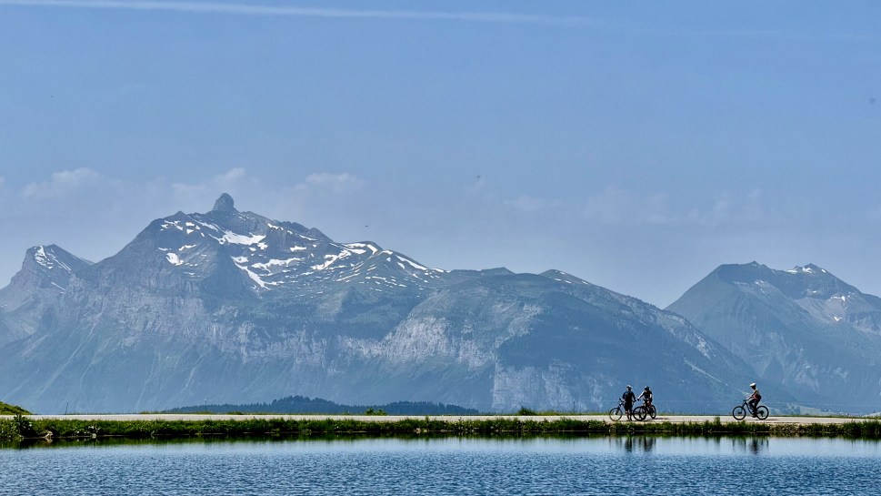 A scenic view showing cyclists riding near a lake with mountains in the background, under a clear blue sky.