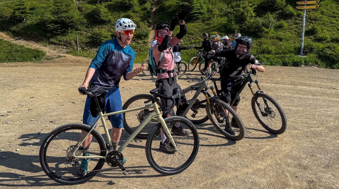 A group of four mountain bikers posing together on a dirt trail, surrounded by greenery and marked trail signs.