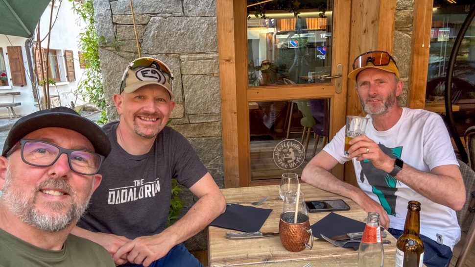 Three friends enjoying drinks at a cafe in Morzine, France, with a rustic stone wall and wooden door in the background.