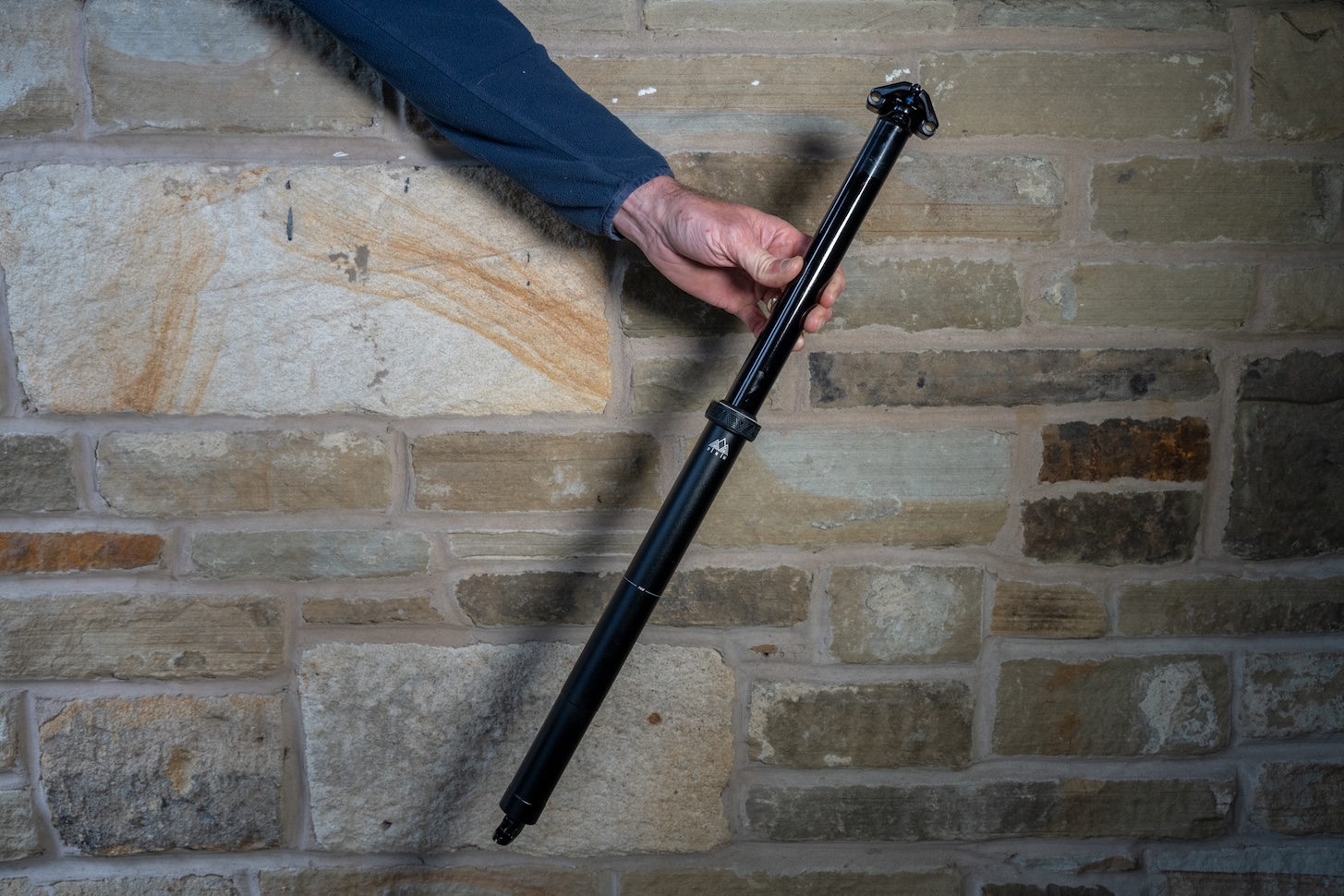 A person holding a black dropper seat post against a stone wall.