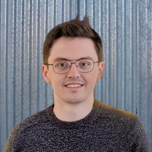 A portrait of a young man with glasses and short hair, smiling against a corrugated metal background.