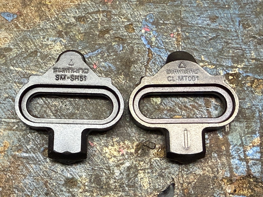Close-up view of two Shimano cleats, labeled SM-SH51 and CL-MT001, placed on a textured surface.