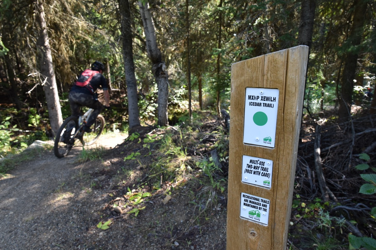 A mountain biker riding on the Cedar Trail in Rossland, BC, with a trail sign indicating the trail name and usage guidelines in a forested area.