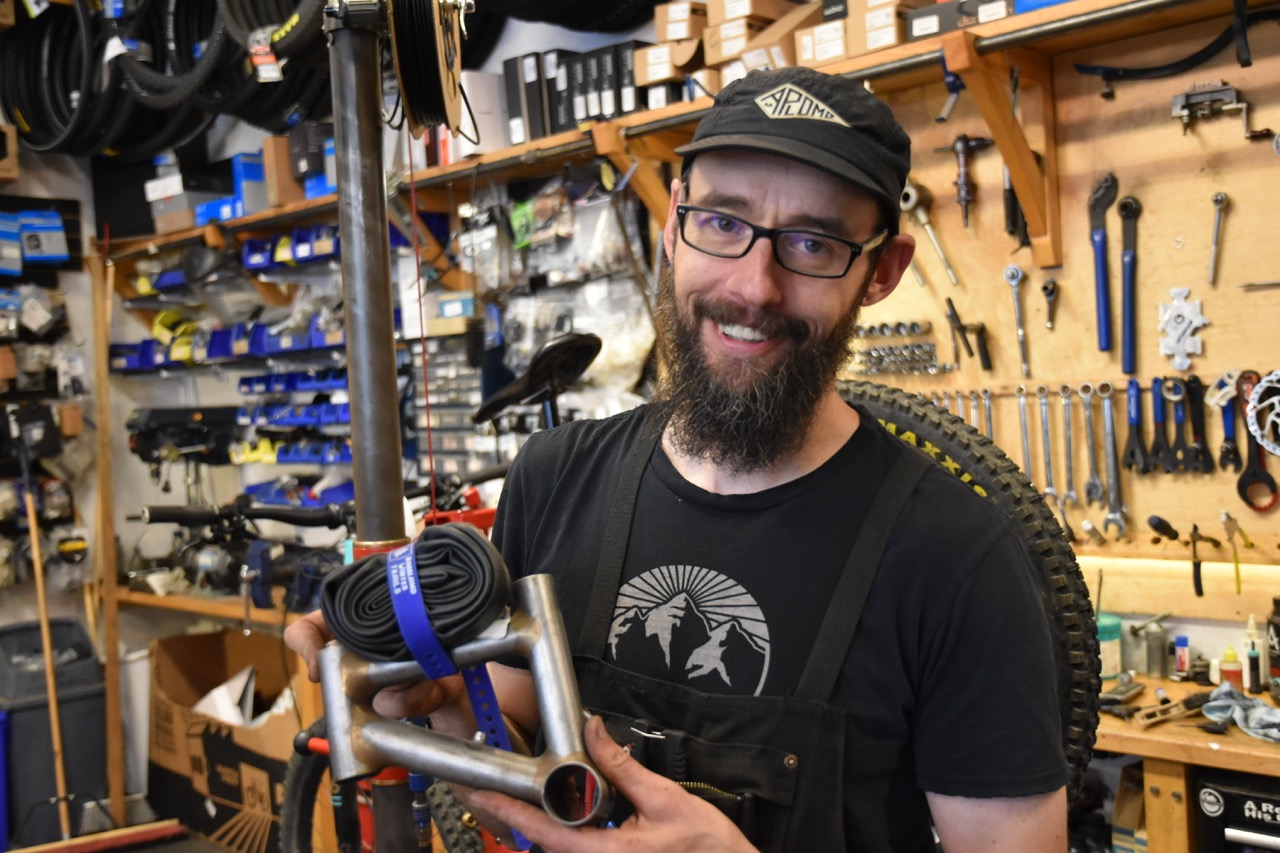 A smiling man holds a bicycle part and tire inner tube in a bike shop filled with tools and bike accessories.
