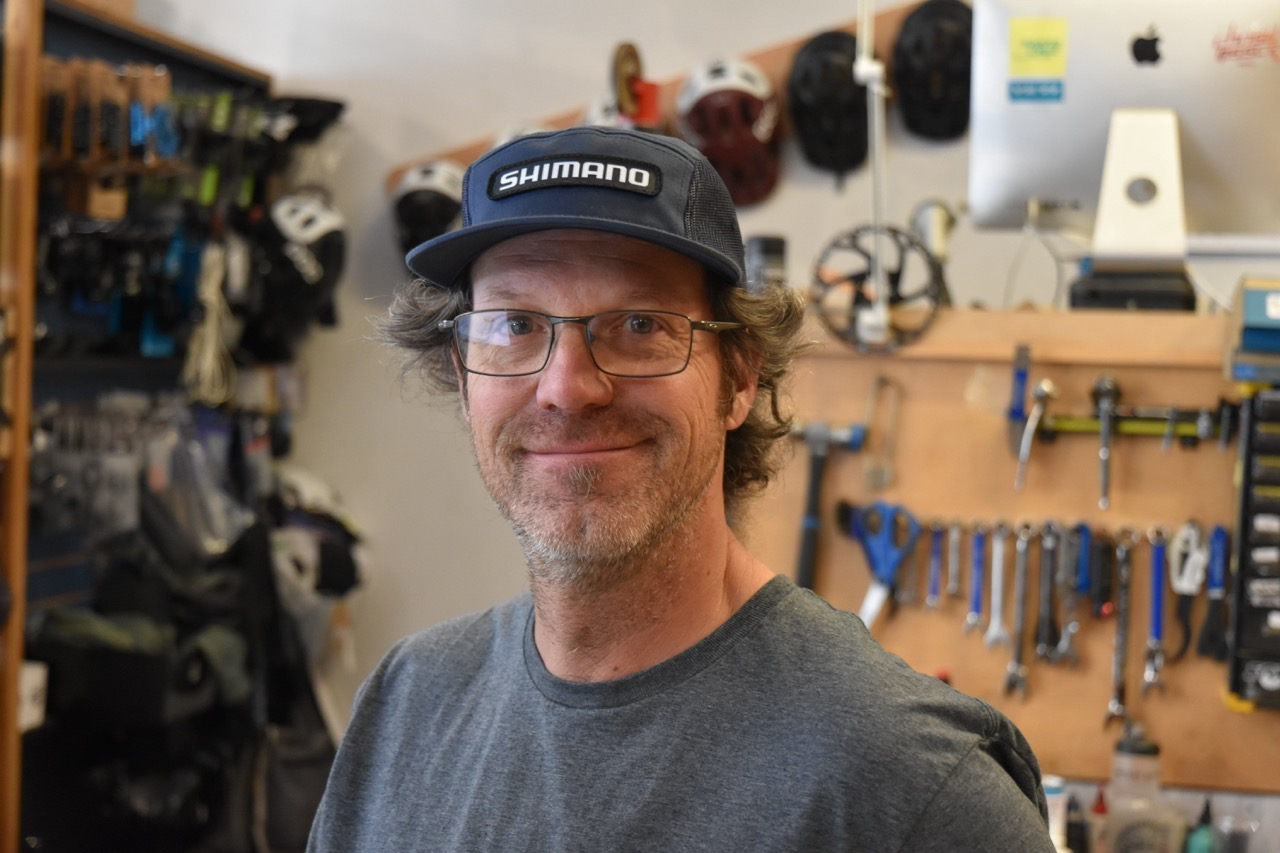 Portrait of a smiling man with glasses and a hat in a bike shop, surrounded by tools and helmets in the background.