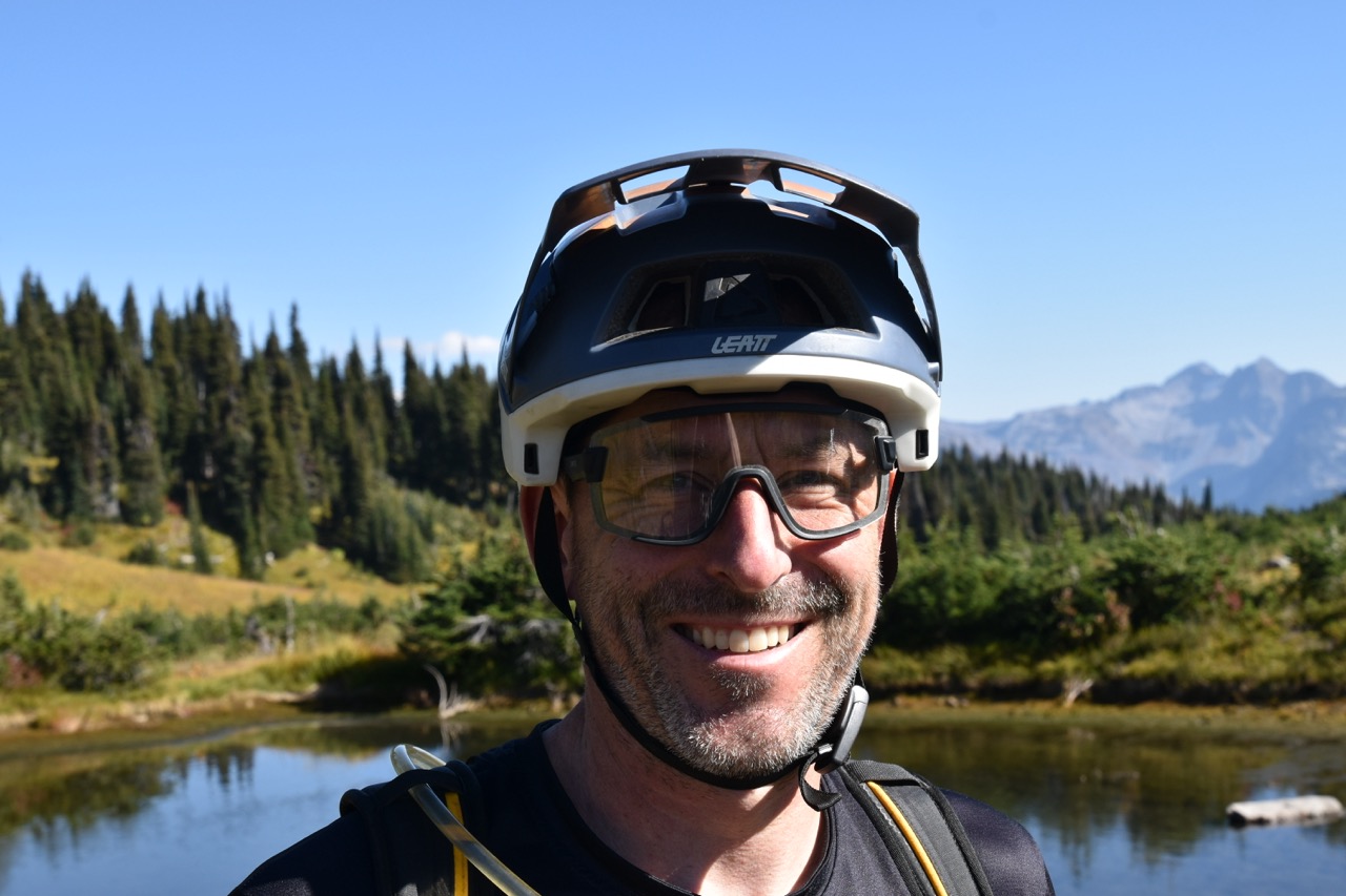 A smiling man wearing a mountain biking helmet and glasses stands in front of a scenic landscape with trees and mountains in the background.