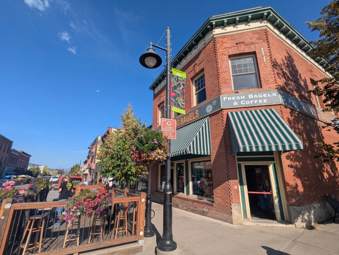 Street view of Big Bang Bagels, a popular cafe and bakery in Fernie, Canada, featuring a brick building, striped awnings, and colorful flower pots.