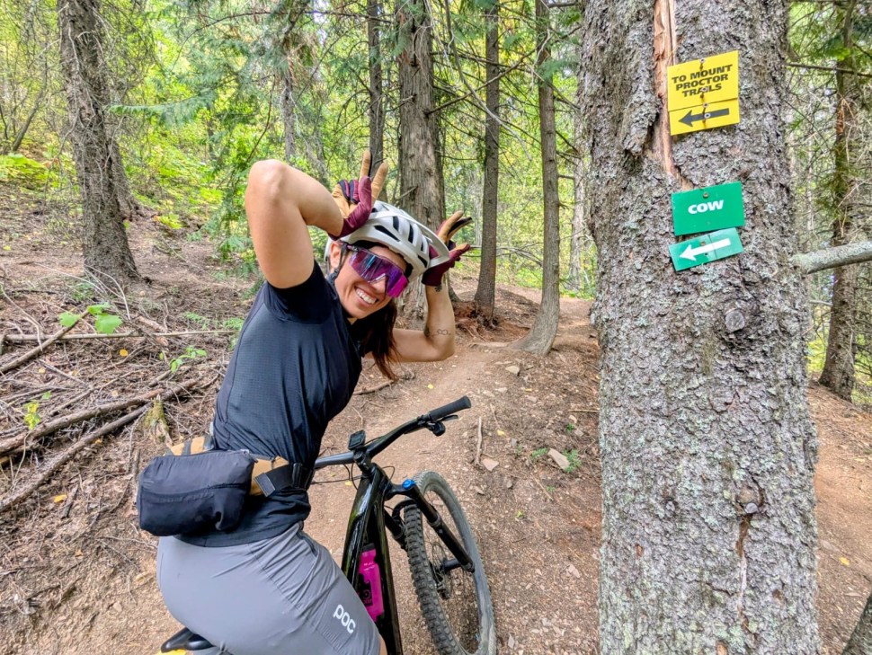 A mountain biker posing playfully on a trail surrounded by trees, with directional signs for Mount Proctor Trails and Cow trail visible nearby.