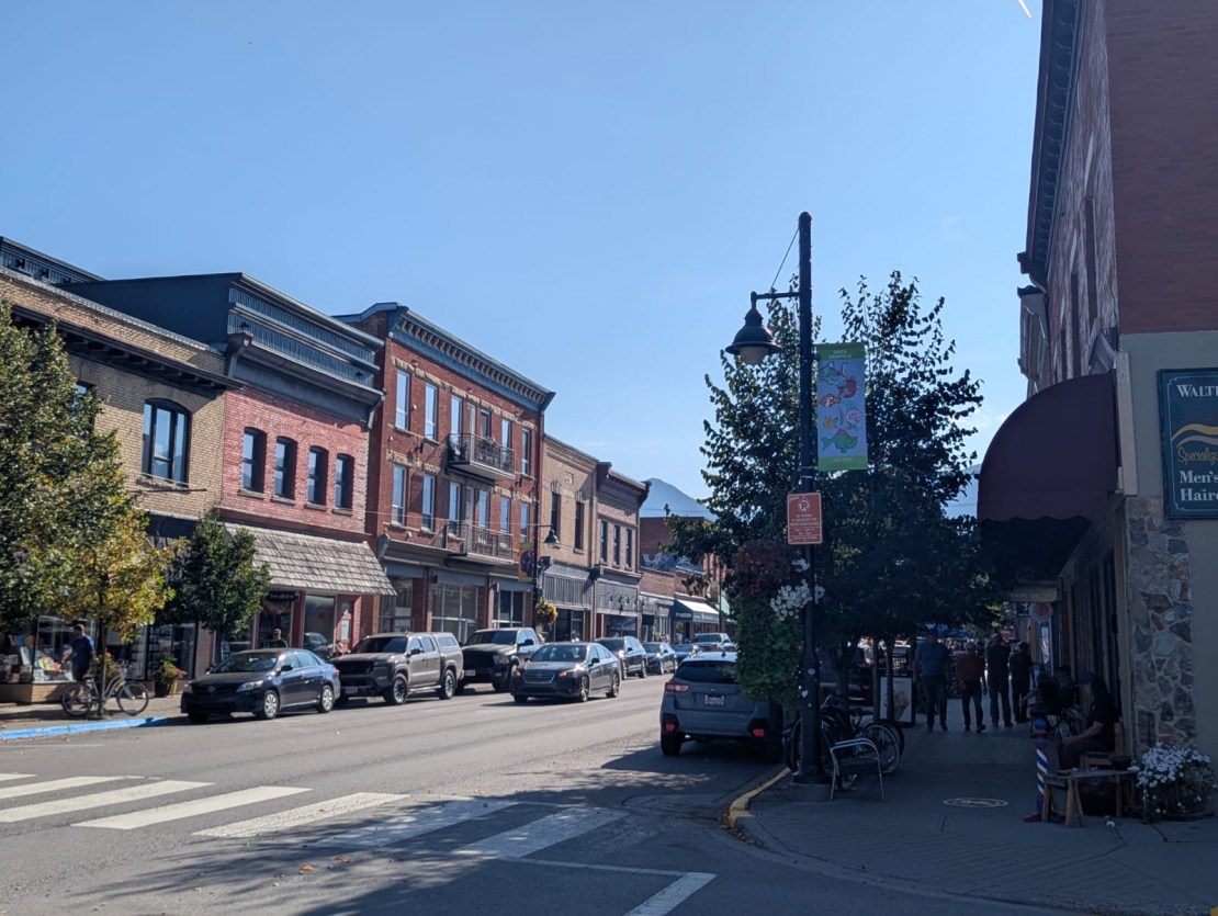A street view of downtown Fernie, Canada, featuring historic brick buildings, parked cars, and people walking along the sidewalk under a clear blue sky.
