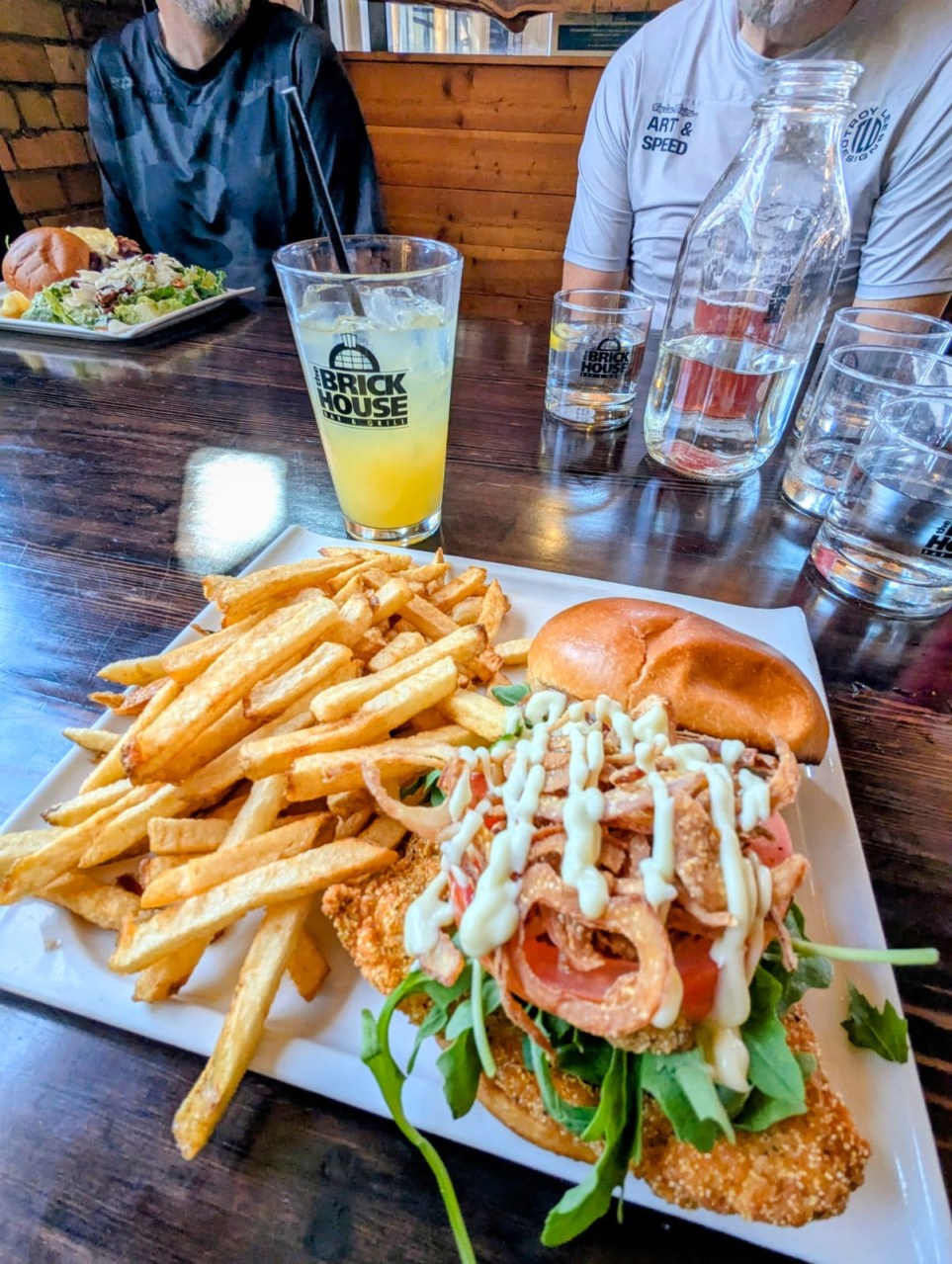 A close-up of a plated meal featuring a fish sandwich topped with greens, tomatoes, and crispy onions, accompanied by a side of golden fries and a beverage served in a glass with a logo.