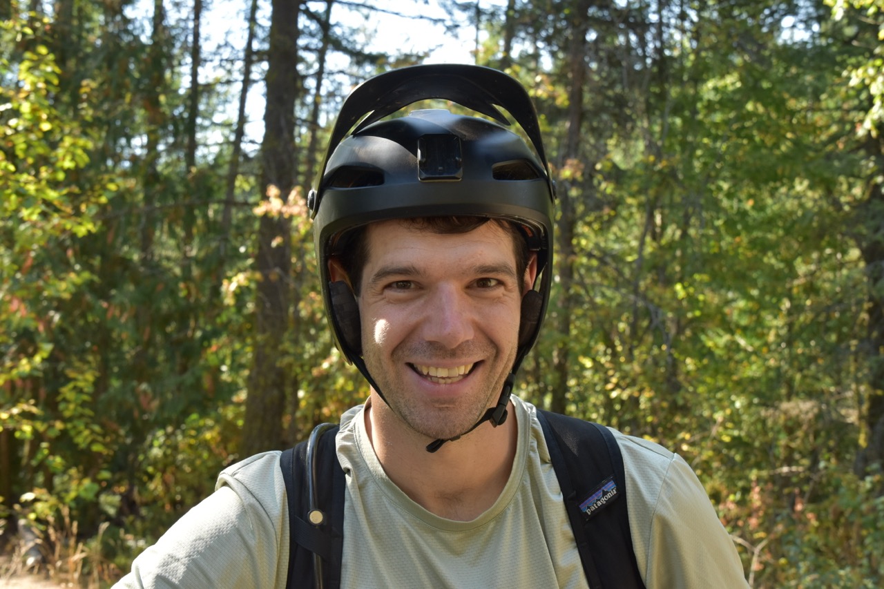 A smiling man wearing a black helmet and a light shirt, standing in a forested area with trees and greenery in the background.
