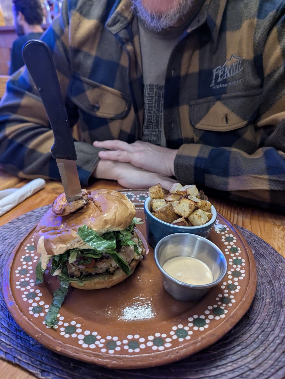 A close-up of a delicious burger topped with lettuce and a slice of pork, served on a wooden plate with a side of crispy potatoes and dipping sauce.